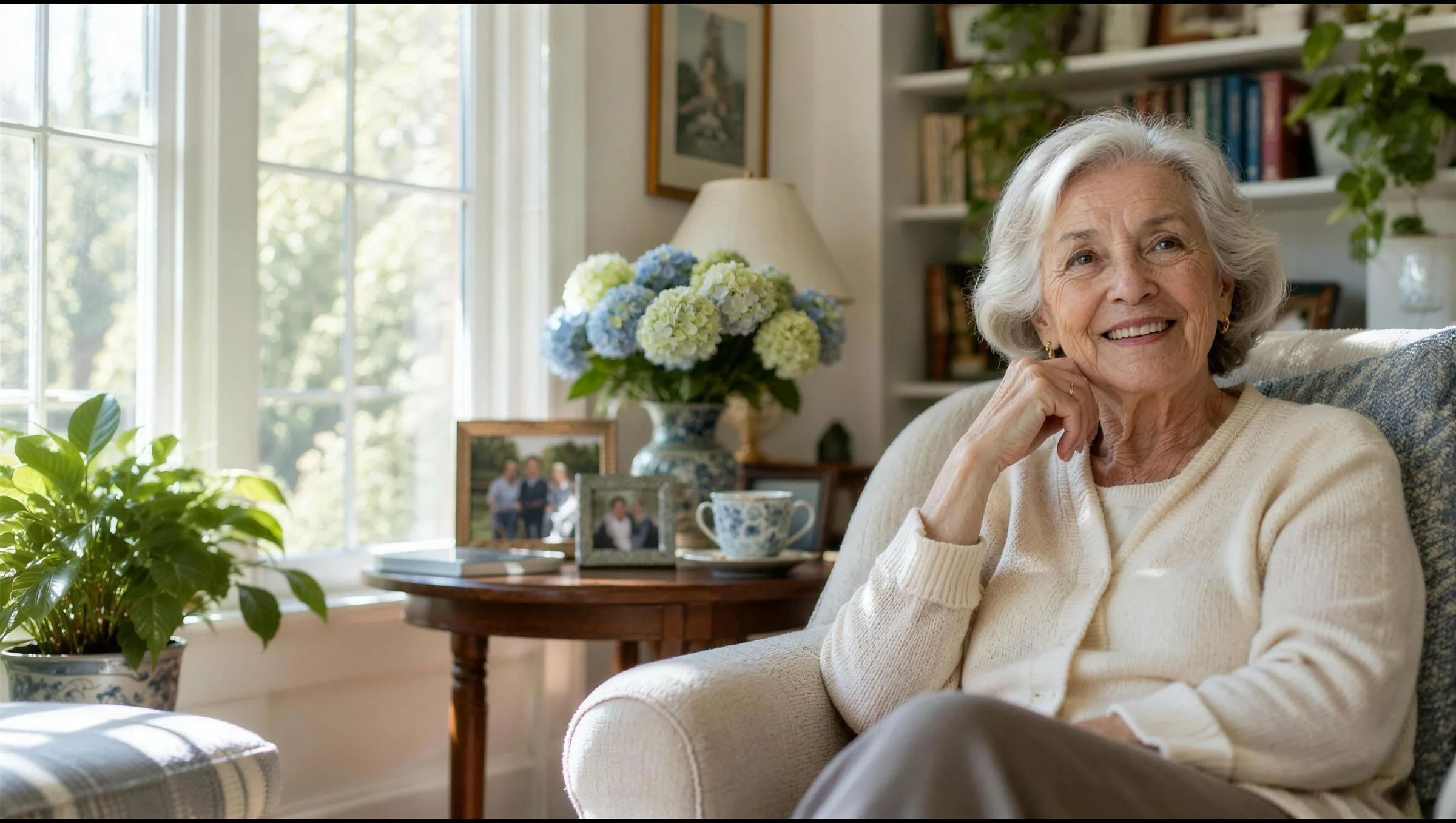 A smiling older woman in a cream cardigan relaxing in an armchair in her sunlit sitting room, hydrangeas and family photos beside her